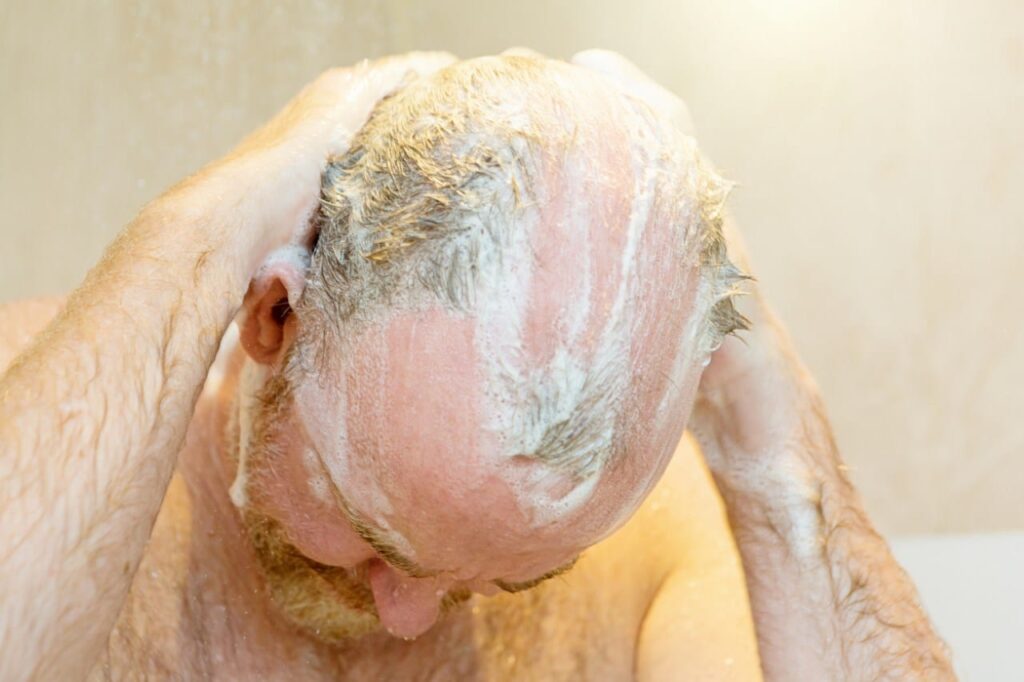 Kel, sakallı bir adam duşta kalan saçlarını şampuanlarken başını öne doğru eğiyor.